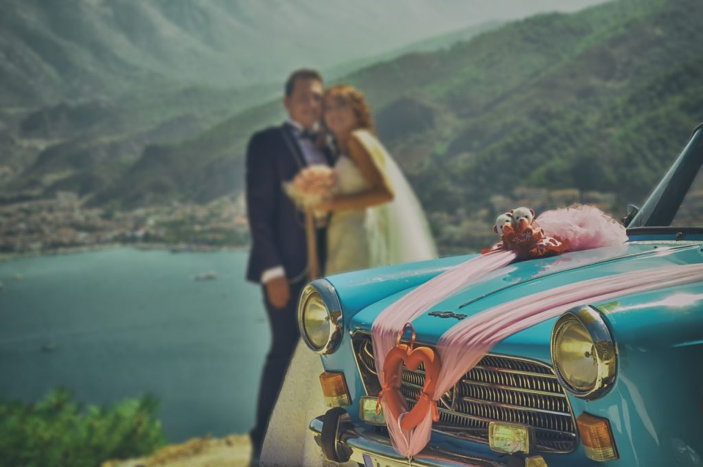 mariés posant pour une photo près d'une voiture bleue décorée avec rubans roses